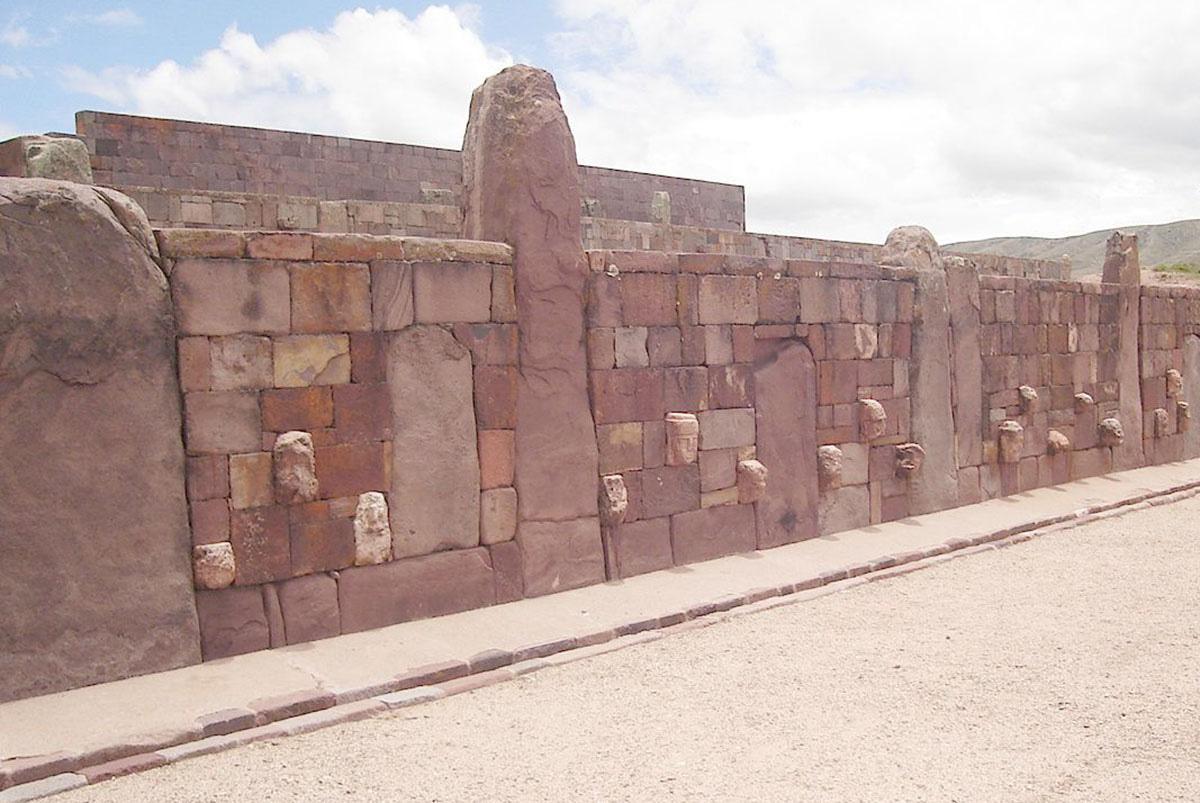 A photo of a long wall is shown along a beige sidewalk with brick edges next to a gravely beige road. A bright cloudy sky shows in the top background. The wall shows square and rectangle shaped bricks of various sizes in a variety of shades of red, pink, and orange divided into sections by four rounded rectangular large stones in pink and gray colors. Some stones project out of the wall along the lower portion in white and beige colors. Three large arched stones can be seen inlaid along three sections of the wall. A large brick structure is seen behind the wall as well as some mountains in the far right distance.