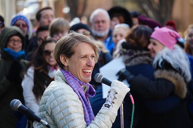 A woman dressed in winter clothing standing holding a micorphone at an outdoor event. A crowd all dressed in winter clohes is standing behind her.