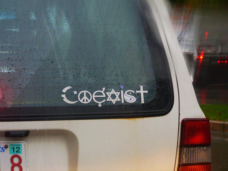 A color photograph of a bumper sticker on the back of a car, spelling out the word “Coexist” using a variety of symbols. For the letter “c,” a crescent moon representing Islam is substituted. For the letter “o,” the peace symbol is substituted. For the letter “e,” a male/female symbol is used. For the letter “x,” the Star of David representing Judaism appears. For the letter “i,” a pagan/Wiccan symbol is used. For the letter “s,” a Chinese yin-yang symbol is substituted. For the letter “t,” a cross representing Christianity is used.