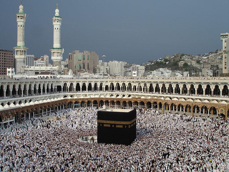 A huge crowd of people stands in an enormous courtyard, with arched walkways tracing the perimeter. In the center of the courtyard is a large rectangular structure, several stories high.