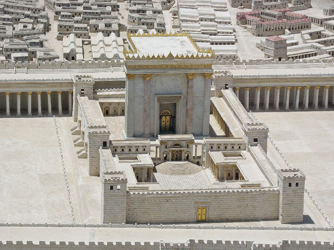 An aerial picture of a large white stone building is shown. The building is tall and marbled gray and white with gold trim across the top and on the roof as well as at the top of the four columns shown on the front. A large framed doorway shows golden doors. A tall white bricked wall surrounds the structure in a rectangle with windowed towers on the corners and in the middle on the long sides. A windowed wall with openings connects the towers on the sides and divides the area in half. In the lower portion the inside wall has openings and a framed opening with gold doors that lead to rounded steps and then to an open area. Buildings hug the inside of the outer walls in this area. A gold door is shown in the lower part of the white brick wall. Buildings can be seen in the background in various shapes and sizes with white, gray, and red roofs. The ground throughout the photo is white stone.