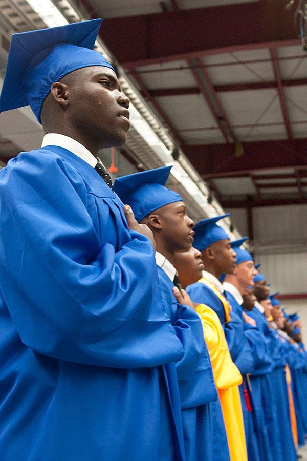 A group of people in graduation caps and gowns stands with their hands over their hearts.