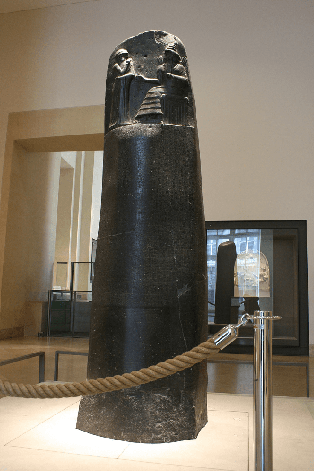 A picture of a tall rounded piece of black wood is shown on a pedestal behind a brown rope. A glass cabinet with a rounded carving shows in the background on the right and a large doorway leading to an open area is seen on the left. The wood is black and shiny, with some drab areas along the bottom. Some cracks are visible throughout. At the top, the front part is carved out showing a person in long robes and a head covering on the left standing handing something to the person siting in a chair on the right. The person in the chair has long ruffled robes on and a tall, pointed, ruffled hat. Lined items project out of his shoulders.