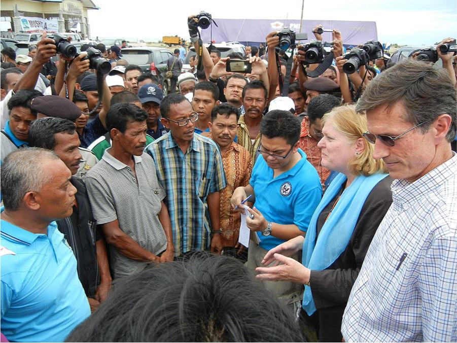 A picture shows a circular group of dark-skinned men standing with a light skinned man and woman on the right side in front of the semi-circle. The dark-skinned men are dressed in various short sleeved printed shirts and short sleeved polos, some have glasses, some wear white hats, and some have facial hair. The woman wears a dark coat, light blue scarf, has blond shoulder length hair, and small earrings. She is holding her hands out in front of her. A man stands to her left in a plaid white and blue shirt, dark glasses, and short, black hair looking down. A man to her right holds a pad of paper and a pen. Toward the middle of the crowd several people hold up cameras, video recorders, and phones. In the background on the left stands a building with a large banner in front, cars can be seen in the far back, and a purple large tarp with a white and yellow flower covers a large rectangle structure toward the middle right of the photo.