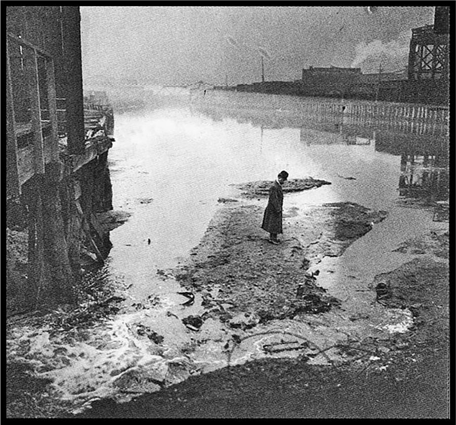 A man in a hat and long coat stands on an island that rises above a pool of liquid waste. Buildings are visible nearby.