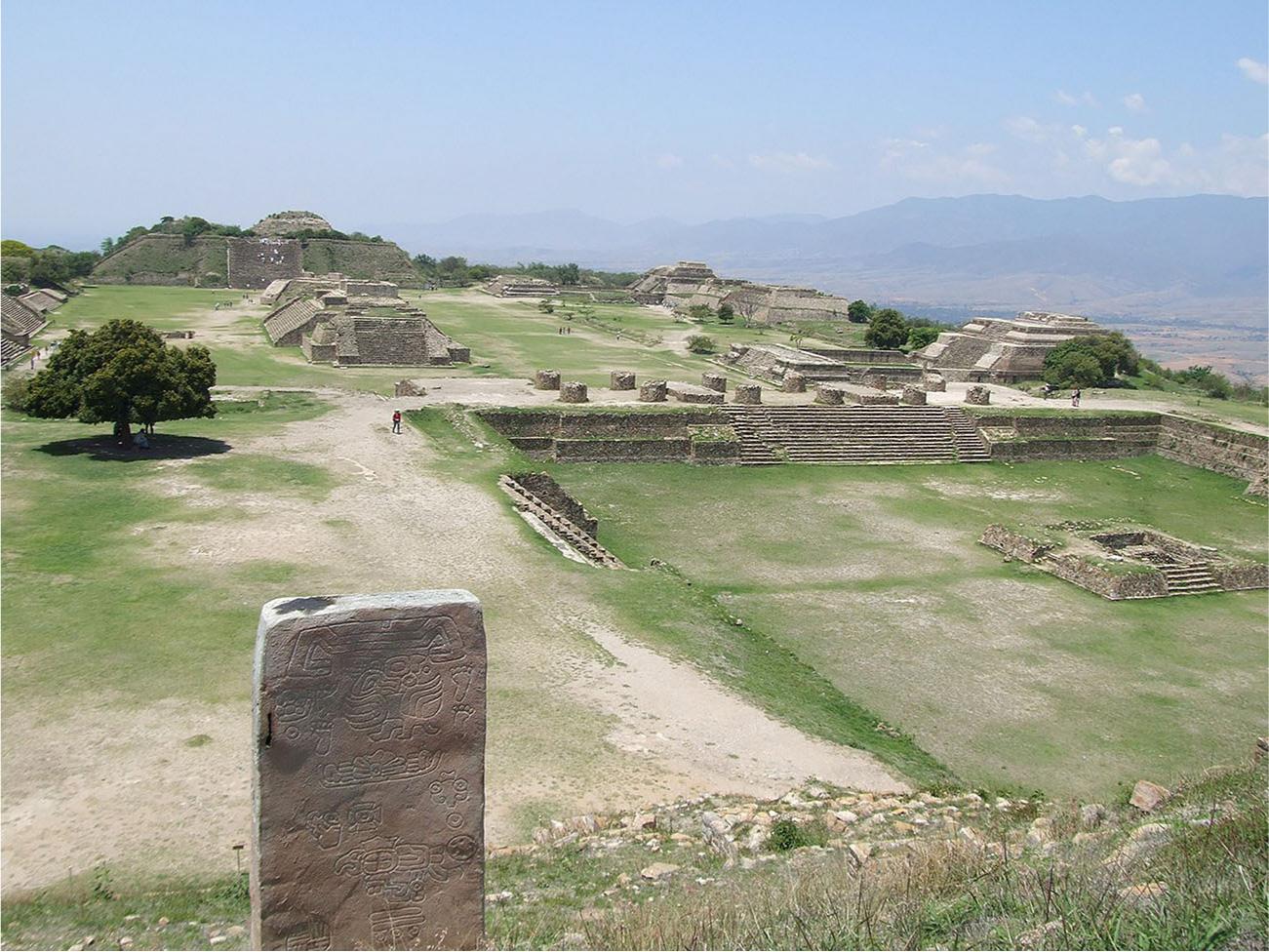 A picture of a large expanse of green land is shown with large mountains in the background. In the forefront a rectangle shaped stone is standing upright with drawings etched all over. To the right is a square section of land sitting lower in the ground, surrounded on the inside by stone bricks and grassy walls. Inside the area in the middle is a square flat section of stones with stairs on one side and grass growing over the rest of the stones. At the back of the lower area are stairs that lead up to higher ground where twelve round stumps of stones stand in two rows. Heading toward the back of the photo there are ruins of many square shaped buildings with stairs leading up to the top. One very tall and large half-triangle structure stands in the back left with stairs seen on the front and mounds of dirt and bushes along the top. Trees are scattered behind the structures and one lone large tree is seen in the middle left of the image.