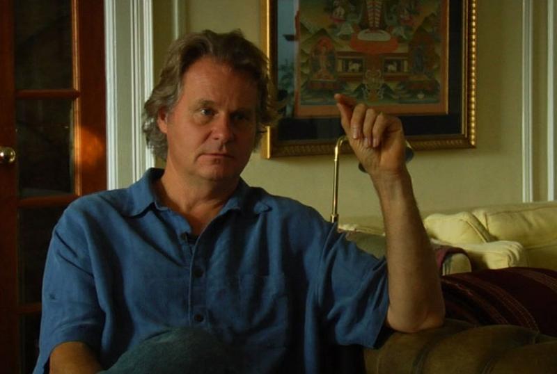 A middle-aged man wearing a short-sleeve button-up shirt sitting on a couch. One arm is raised with his elbow resting on the top of the couch.