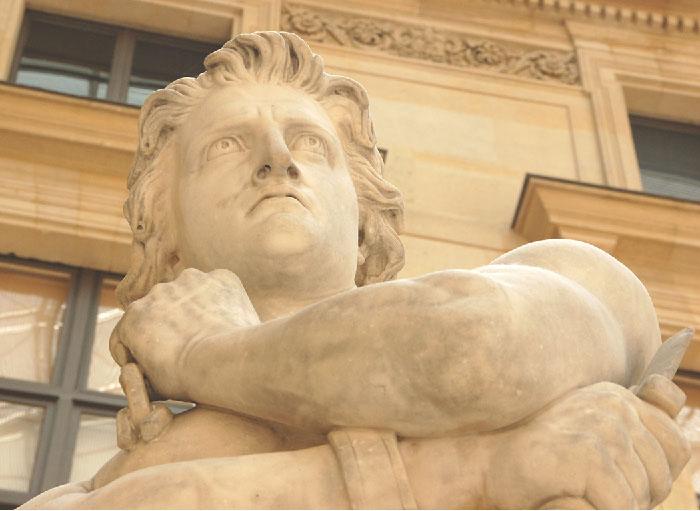 An image of a photograph of a beige stone statue is shown with beige buildings with windows in the background. The view is from the bottom looking up. The man’s face is shown with his arms crossed in front of his chest. He has wavy hair, large half-circle eyes, flaring nostrils, and a downturned mouth. One of his wrists shows a bracelet and he holds a knife in his right hand.