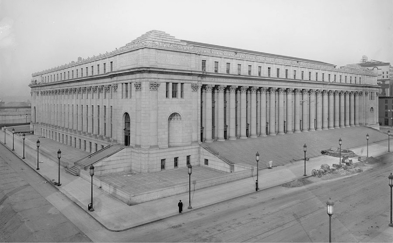 A black and white picture of the corner of a large stone building is shown. Tall columns decorated at the top line the outside of the building on the two sides shown. Rectangular windows line the top of the building and the corner is bricked with an inscription over the colonnade on both sides of the corner. An arched doorway is shown on the left side with stairs leading up to it from the sidewalk. A recessed arched alcove is seen on the other side of the corner. Stairs run the length of the building on the right side. Lampposts line the street on both sides and a road is seen in the forefront on both sides of the building. Other buildings can be seen in the background and a lone figure stands at the corner in a long black coat and hat. Some wheels and piles of dirt can be seen on the right side of the building in front of the stairs.