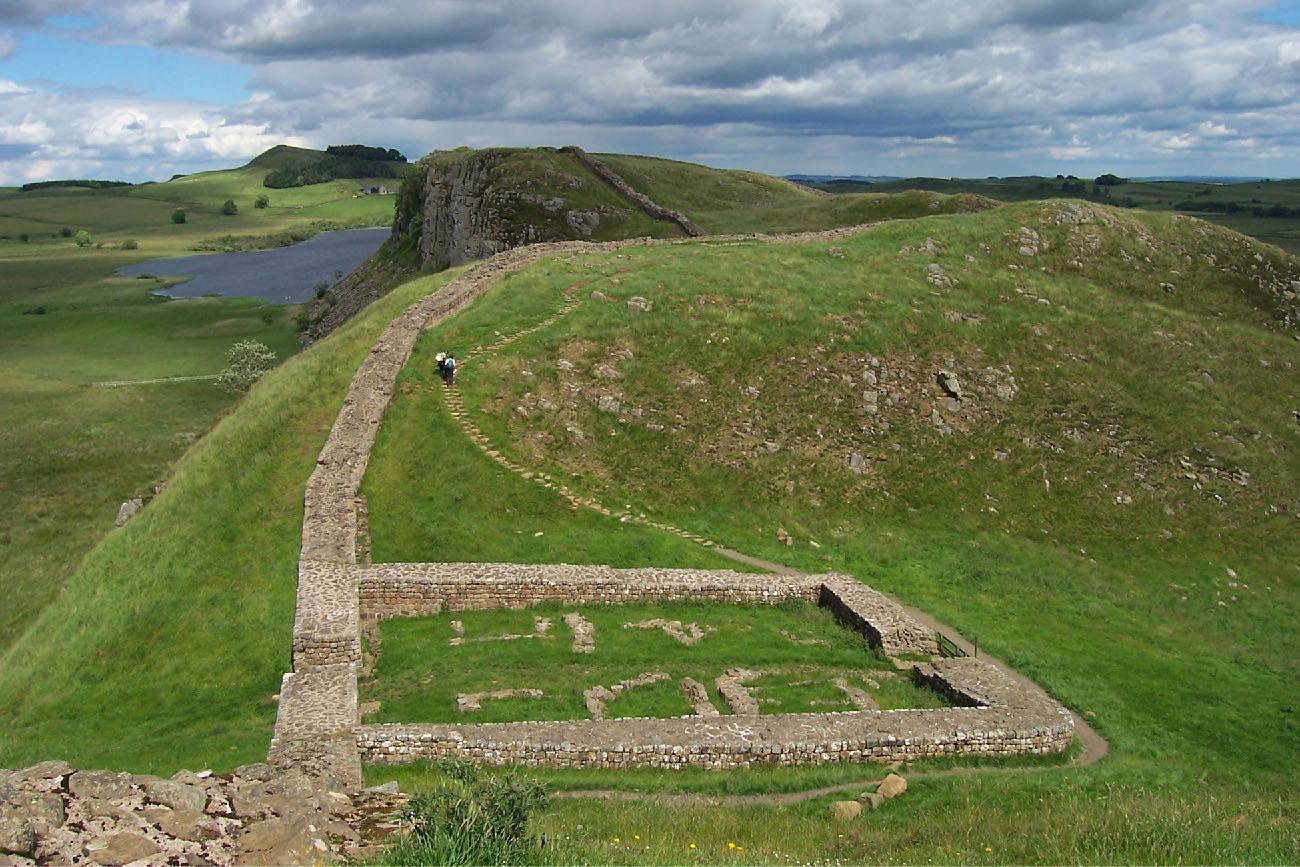 An image of a photograph of a green countryside is seen with a gray and white cloudy blue sky in the background. In the forefront, a low brick outline of a square area can be seen with some small squares lined with stones on the inside. Green grass fills in the rest of the area. Connecting to the square, a low stone wall continues in a curved line to the back right of the image and over rolling green hills to the left. A flat dirt path goes around the square wall and continues as a thin stone path running along the low wall over the first green hill. Two figures are seen walking on the path dressed in white and black clothing. In the background a pond can be seen and sparse trees and bushes over green terrain.