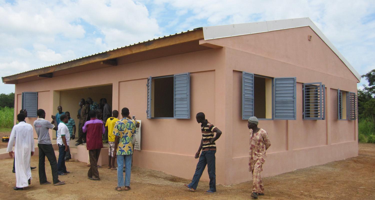 Group of African men waiting outside of a one-story building. The building has many open windows with shutters. The men are wearing both traditional and western clothing.