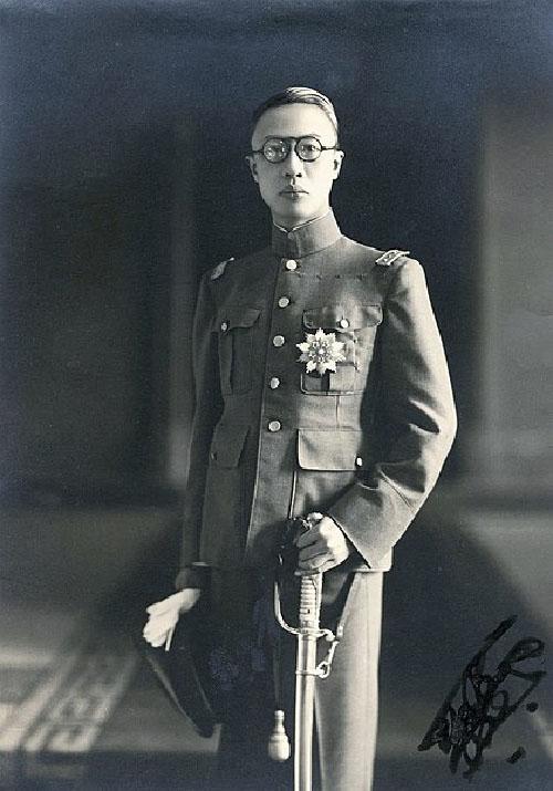 A man wears a military style uniform and holds a sword. He stares directly into the camera.