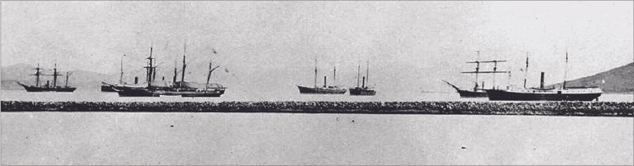 A photograph shows seven ships with tall masts in water. They have black hulls and no sails up. A stone breakwater is shown in the foreground and the background shows small hills.