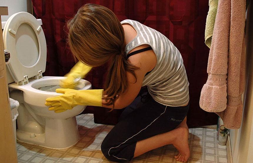 A woman is shown kneeling on bathroom floor scrubbing toilet.