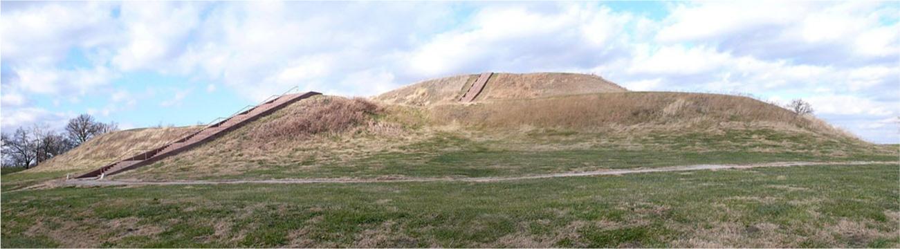 A photo is shown of a large, two-tiered mound with brown grass growing on it and a staircase leading from the bottom left to the top with railing on the sides. The grass at the bottom of the mound is green and a path can be seen in front of the mound running the length of the photo. The blue sky can be seen in the background with white clouds and trees are seen in the far left.