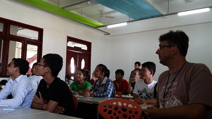 Teenagers or young adults sit at tables in a classroom. They are all dressed casually, and many are smiling or laughing.