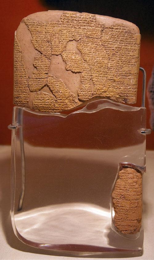 A picture of two pieces of a brown stone tablet are shown on a black, maroon, and beige background held up by a wire stand. Fragments of the top half of the stone and the small piece of the right side have script on them while the pieces that are chipped off show the smooth stone underneath. Clear thick glass is shown replacing the pieces of the stone that are missing in the bottom half.