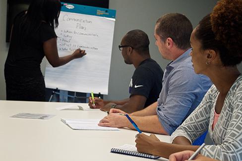 A photograph shows several people sitting at a table and writing on notepads, as a person in the front of the room writes on a large tablet.