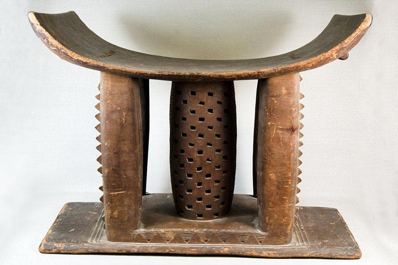 A color photograph of an ornately carved wooden stool on a base. It is connected to the base by four thick posts on each corner and a thicker carved post in the center. The seat is curved, higher on the outer edges. The overall look of the stool is solid and strong.