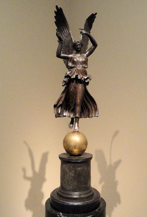 A picture of a bronze-colored statue of a winged female is shown on a round, tiered pedestal. Her large feathered wings extend above her head and she wears a long sleeveless dress with ruffles swirling below the waist. Her arms extend above her head and she has short hair. Necklaces hang from her neck and one of her bare feet stands on a golden sphere. Her other bare foot extends out toward the back. Shadows form on both sides of the pedestal from her figure on the yellow ombre walls behind the statue.