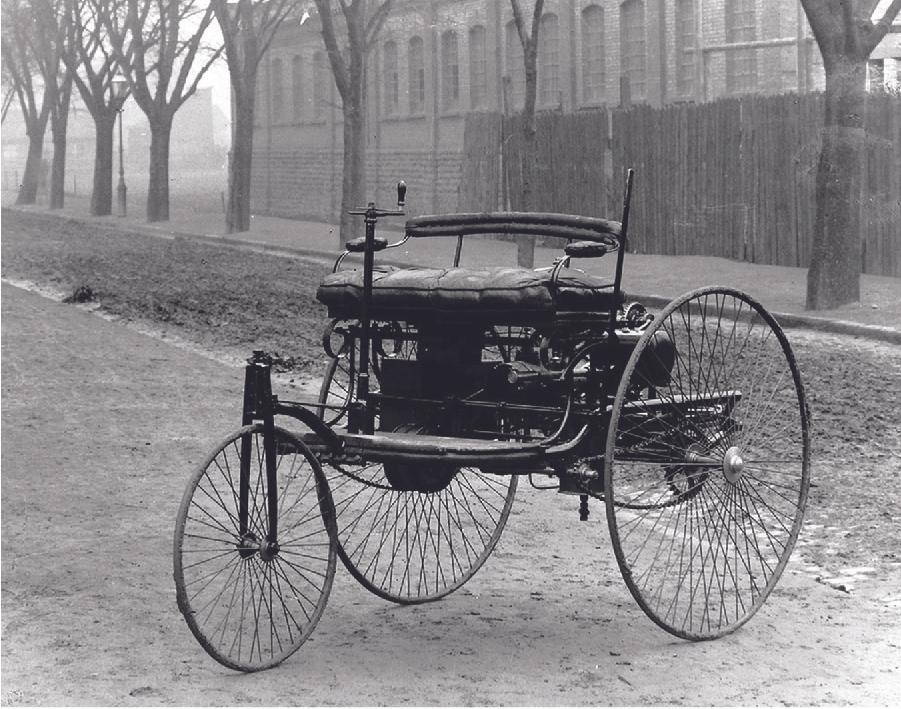 An old photograph shows an automobile. It has two tall thin wheels in the back and one smaller thin wheel at the front with many very thin spokes on all three. A floor sits high off the ground and a two-person cushioned seat with a back rail and side arms sits on that floor. A crank is on the car’s right side in front of the seat. There is gadgetry between the seat and the floor. The car is on a road with a building, a wooden fence, and trees shown in the background.