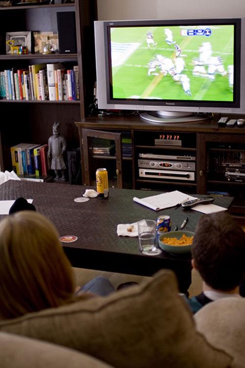 A boy and girl are shown from behind watching a football game on television. A coffee table sits between them and the television, and a bookshelf is beside the TV.