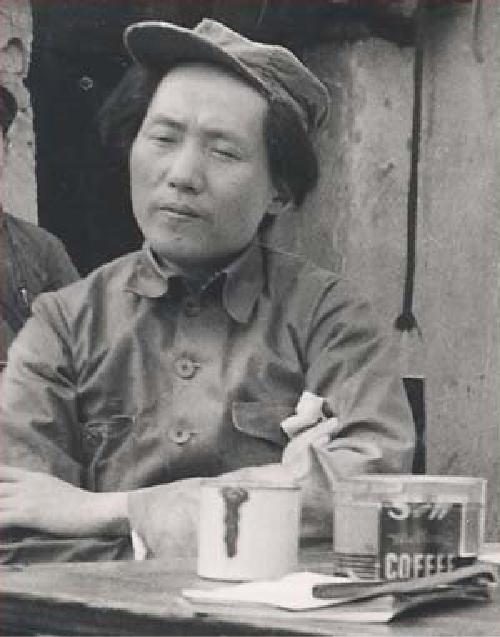 A man wears a military style uniform. A coffee cup and coffee can sit on the table in front of him.