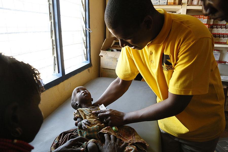 A person holds a baby who appears to be about two years old. The baby is on an examination table. In the background are boxes of medicine.