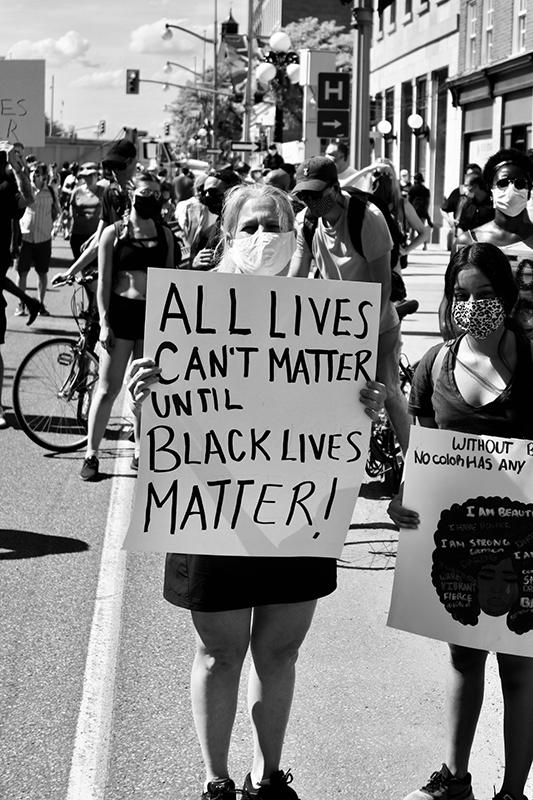 A woman stands in the street holding a poster with the words “All Lives Can’t Matter Until Black Lives Matter” written on it.