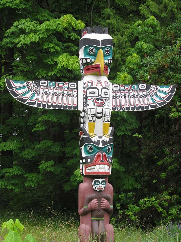 Totem pole with two figures stacked one atop the other. The top figure is birdlike, with large extended wings. The bottom figure holds a smaller figure against its chest.