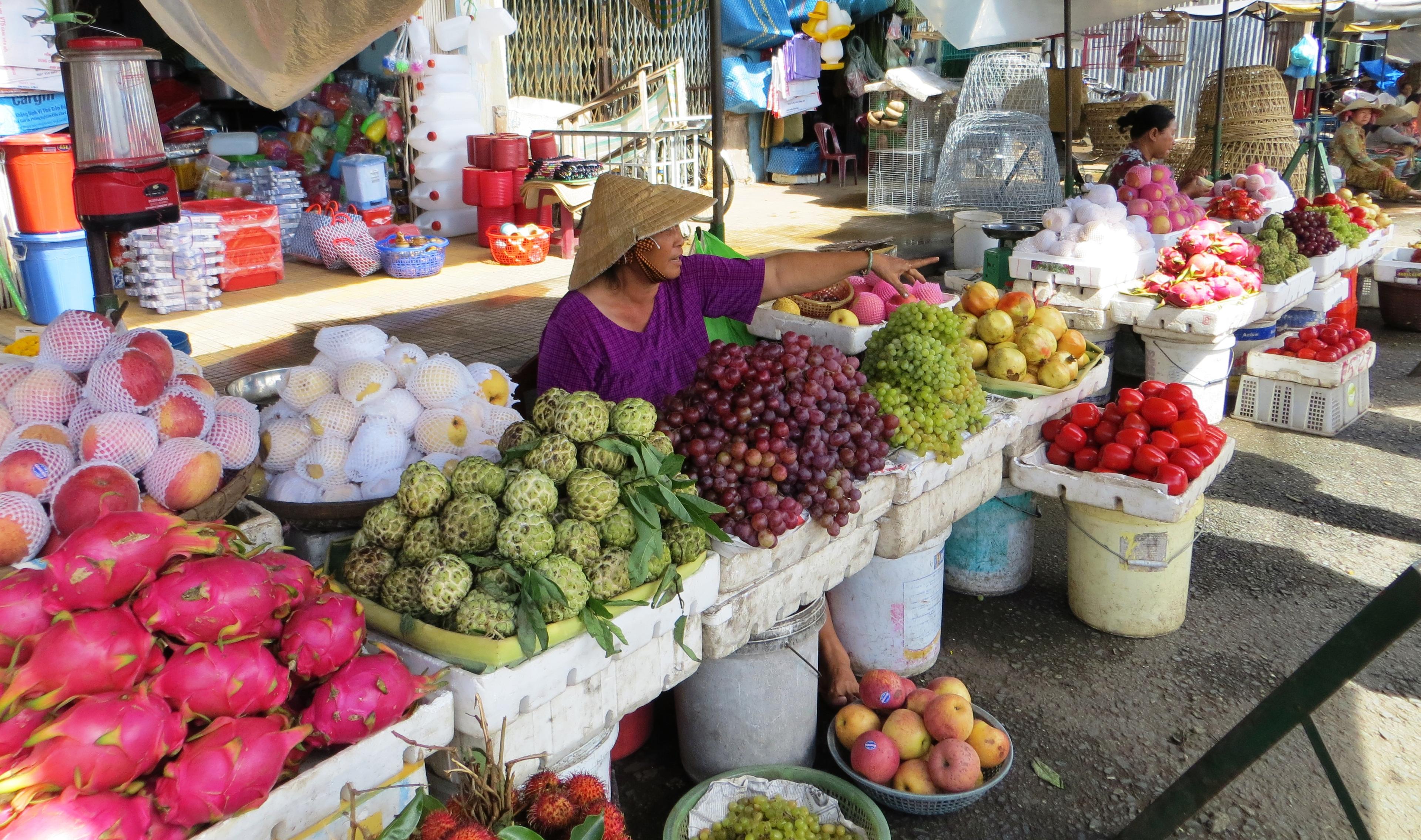 A woman wearing a cone-shaped sunhat stands behind tables piled high with fruits and vegetables. Some of the produce will be familiar to a Western viewer, such as grapes and apples. Other items, such as a stack of bright pink oblong-shaped fruits, are not commonly eaten in the United States.