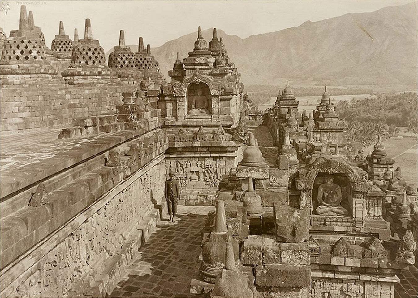 An image of a sepia colored photograph is shown. The far right background shows tall mountains and areas of trees, water, and flat grassy areas show in front of the mountains on the right of the image. At the top left, eight mound-like structures (stupas) are shown sitting atop square bricked pedestals at varying levels. A thin vertical spire shows at the top and four rows of diamond shaped perforated openings line the mound. To the right, more solid stupas line the top of a structure with an arc shaped opening with a sitting statue inside. The archway is decorated with carvings. To the left of the structure on the same level as the stupas, a broken wall of bricks shows with the head of a statue showing. The next level down shows an uneven bricked walkway that extends out to the back of the massive structure. Lighter colored walls run along the walkway made of bricks and carvings show within those bricks. A figure stands along this wall under the sitting statue above in a dark buttoned jacket and pants that stop below the knee. He wears a dark cap and looks down. To the right of the walkway in the forefront, another sitting statue in an arched opening sits with legs crossed and hands resting in his lap. The statue is very worn and the stones above the arch are broken and missing pieces. Another walkway sits to the lower right of this structure with walls running along with carvings as described earlier. Along the right side, walls run with solid stupas lining the tops of varying levels.