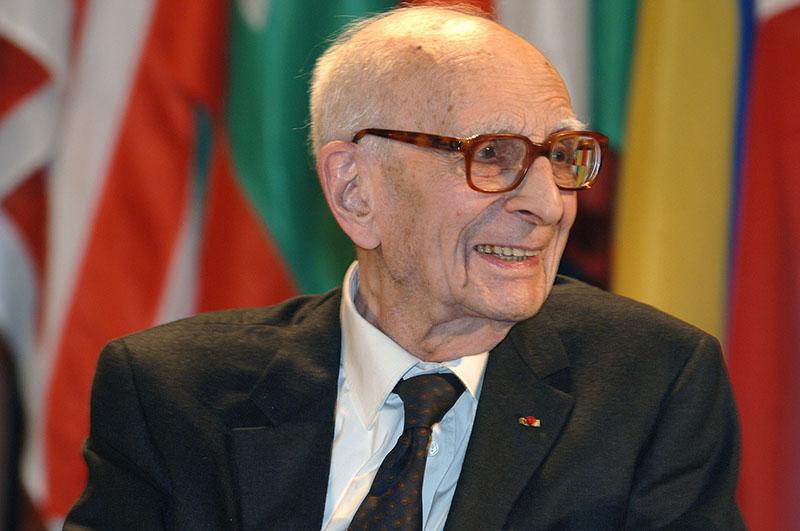 Contemporary photograph of an elderly man wearing large glasses and a black suit jacket.