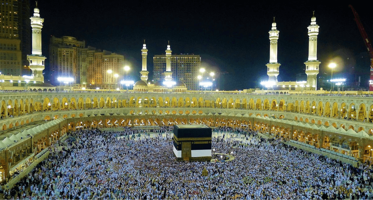 A picture of a very large gathering area is shown. The open area inside is filled with people, most of them wearing white head coverings. In the center of the people stands a short square building that is covered in black. The perimeter of the area is two tiered with arched openings all around and lights and people showing from within. Lights show along the top and on the back wall three domed projections stand along the top of the wall. Five tall slim towers also rise up from the top wall and are lit up and carved with designs. In the background a dark sky is seen and a city with tall lit up buildings.