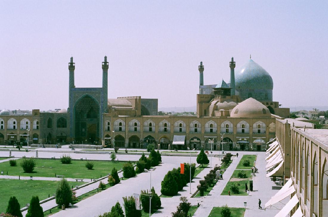 A photograph of two long building in a “L” shape is shown. Starting on the left, a long building is shown with a large arched opening and thin towers on either side. The building has many windows on two floors and runs the length of the photograph. The right side of the building shows domes and towers rising form the second floor. Along the right side of the photograph another long building is shown with two floors, windows on both levels and awnings coming off of the windows on the lower floor. In front of both buildings are trees, grass, sidewalks, light poles, shrubs, people, and roads.