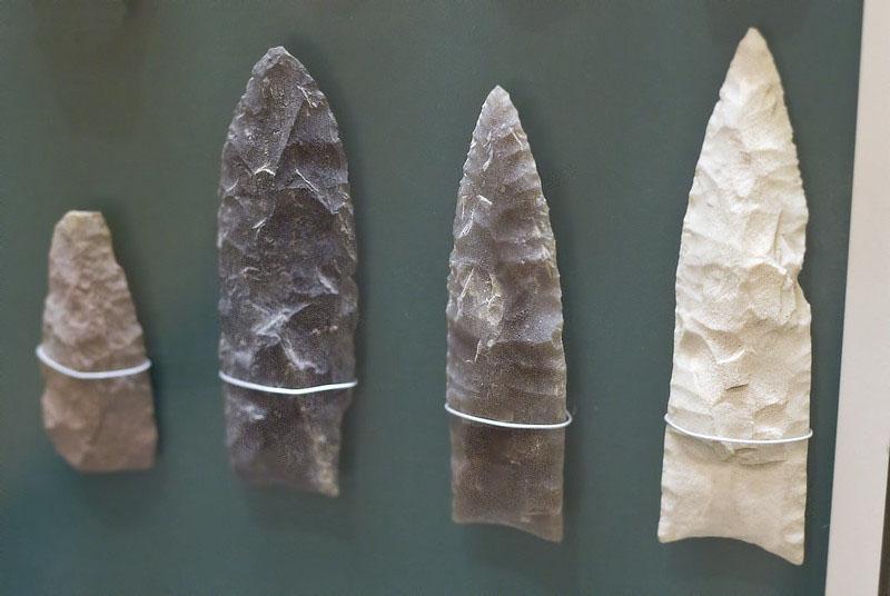 A color photograph of four shaped stones mounted to a display board. They are of various tones, one bright white and the others darker shades. Each is vaguely rectangular at the bottom and shaped into a blunt point at the top. All four stones have an uneven surface, with evidence of chips having been removed.