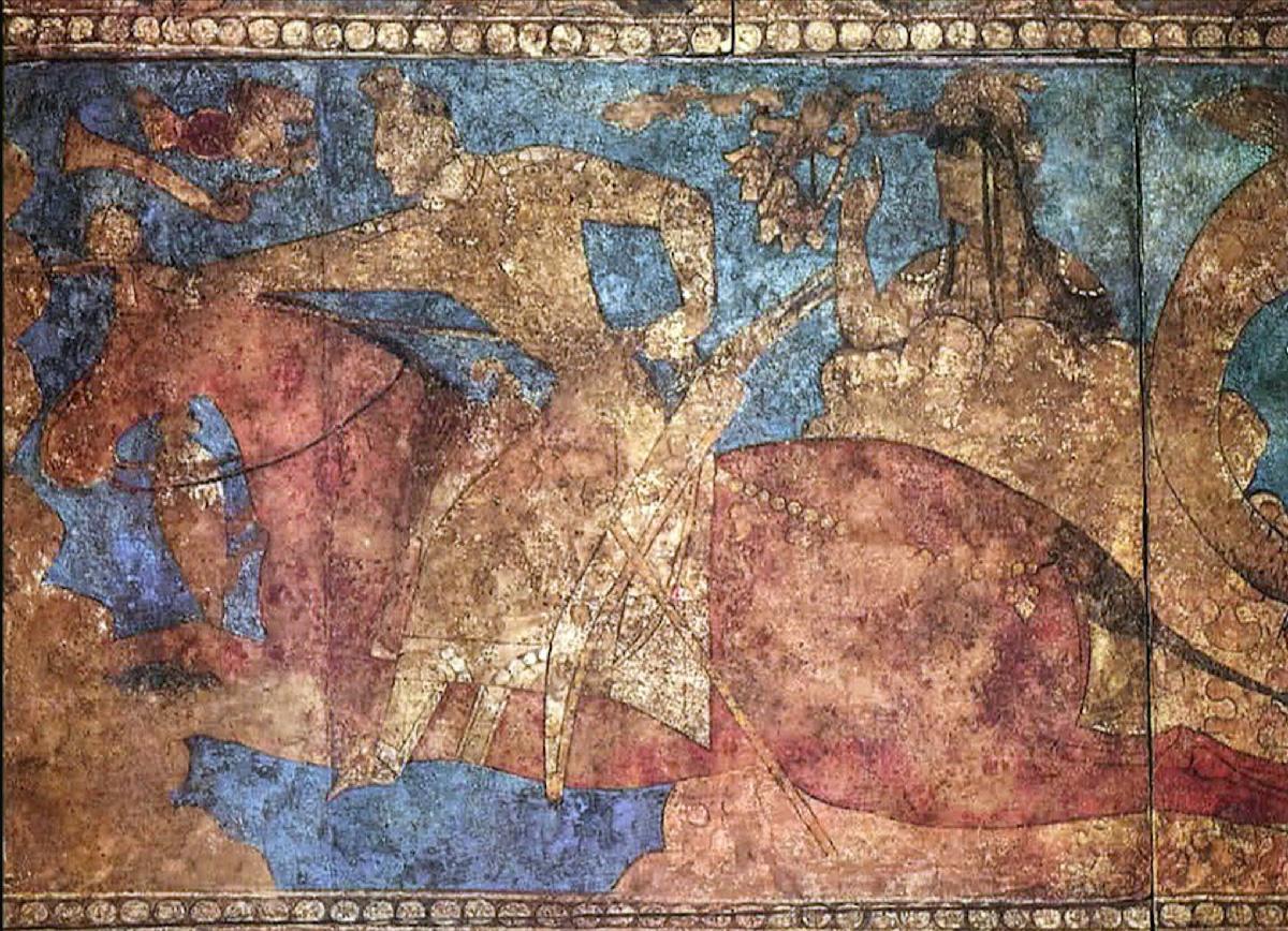 An image of a wall painting is shown. The top and bottom borders are a single row of gold circles. In the image, a gold figure in a long robe and no facial features is seen atop a red horse with no features. He has a sheathed sword at this belt and he holds the reins in his left hand. Behind him is a figure with long black hair, an intricate gold headdress, jewelry, and no facial features. The figure stands behind a gold object and a blue sky shows behind all of them. Obscure objects are seen in the top left of the image.