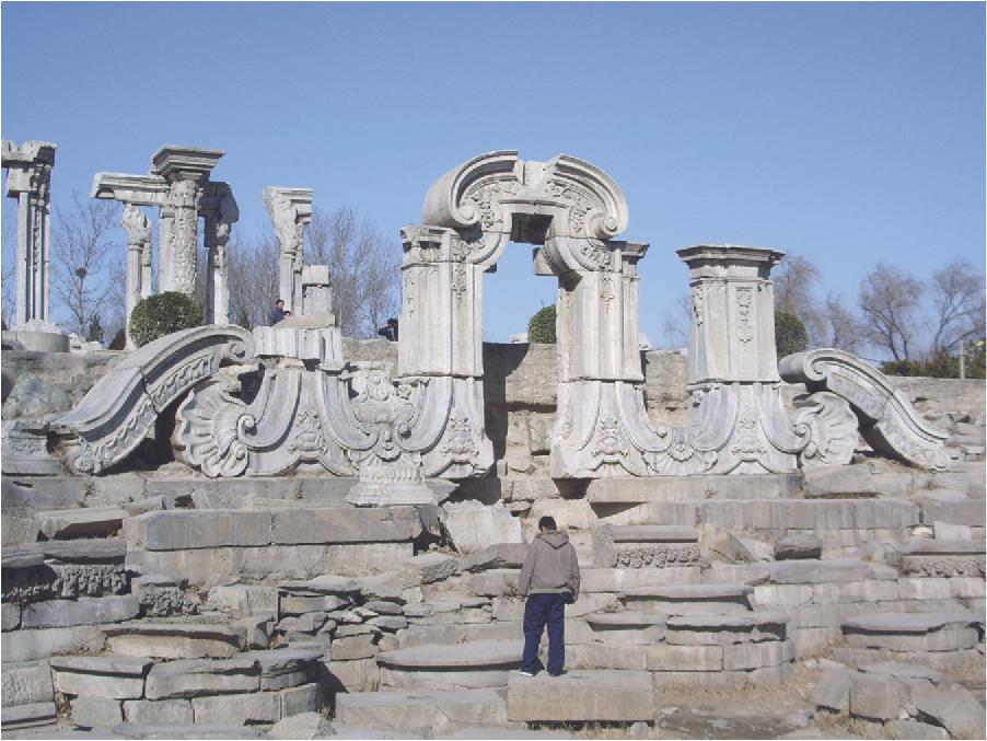 A photograph shows a very detailed white stone archway surrounded by collapsed and broken archway pieces. In the back left of the picture are five tall broken columns that are richly engraved. Trees run along the background.