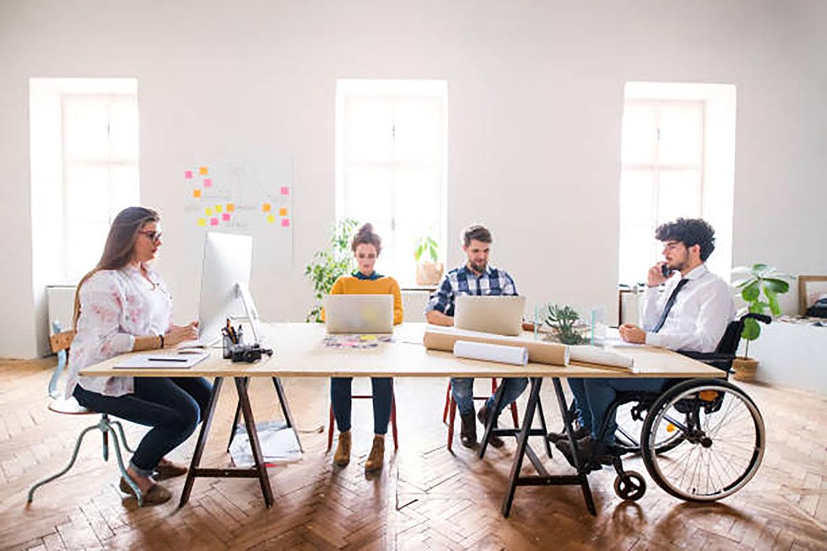 Four people sit around a large table on which are several office supplies and mailing tubes. Three are using computers; the fourth is using a phone. One person is using a wheelchair.