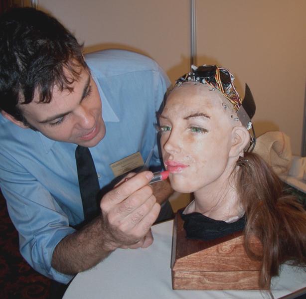 A very life-like head with wires coming out of the top emerges from a block of wood. A person leans over and applies lipstick to the lips of the head.