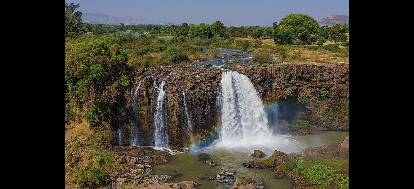 A picture of a landscape with a waterfall is shown. The upper portion shows blue skies with mountains in the far distance on the right and left. Trees, grass, yellow fields, and a snaking waterway are at the top of the hill. The waterway spills down a rocky mountainside in a large waterfall on the right and three small ones on the left into a rocky river with dark colored water. A rainbow forms to the right and left of the large waterfall.