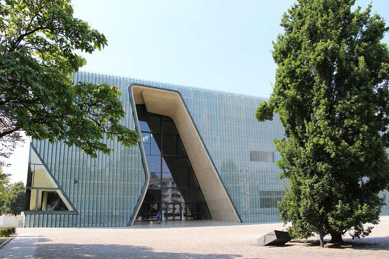 A color photograph of a large building constructed in a modernist style. The building is vaguely rectangular and covered in smooth and shiny opaque glass panels. In two places, large geometric cut-outs in the opaque covering reveal clear glass, giving a glimpse of the interior.