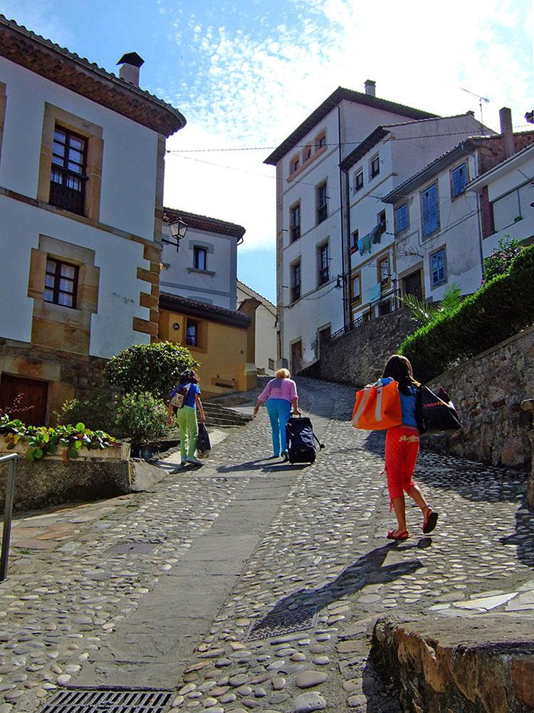 Three people carry luggage up a steep hill paved with cobblestones.