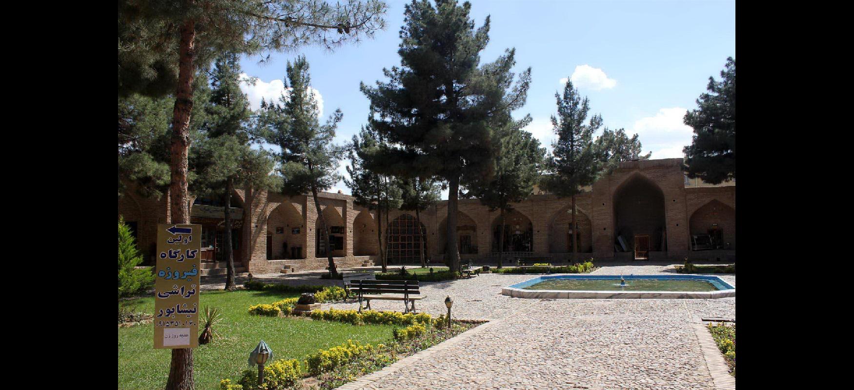 In the background of this picture, a light brown brick building is seen with ten pointed arched openings, one taller than the others on the right. Tall trees with long trunks are in front of the building. A tan, pebbled walkway is seen on the right surrounding a square pool of water with a small sprayer in the middle. To the left, green grass is bordered by yellow flowers with small lanterns at the corners. A bench is seen on the path and a sign with Iranian writing attached to a tree is located in the left forefront.