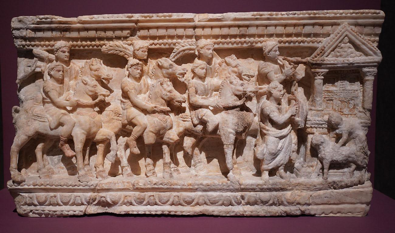 An image of a stone carving is shown on a maroon colored background. The stone is off-white and cracked in places. Designs made up of squares, circles, and “U” shapes run across the top and bottom of the carving. At the right an altar is seen with a carved bottom, two columns in the middle with a highly etched box in between the columns, and a triangle top with décor on the perimeter and the carving of a faded figure in the middle. A small person in a shirt is shown with no facial features in front of the altar, bending over and holding on to a small furry looking animal on four legs with no snout visible. To the left of the altar stand two figures with short hair, robes, holding items in their hands. The one in front stands lower than the figure behind who holds a “V” shaped object to his mouth. To the left are seen three groups of two figures, all on horseback. The horses all have decorative reins and saddles and the figures in front are shown lower than the riders in the back. The figures wear robes and shoes with lariats around their heads. The first rider in front is missing a foot. The first two figures in the back have large feathers on their shoulders while the last rider in the back holds a long, round object over his shoulder.