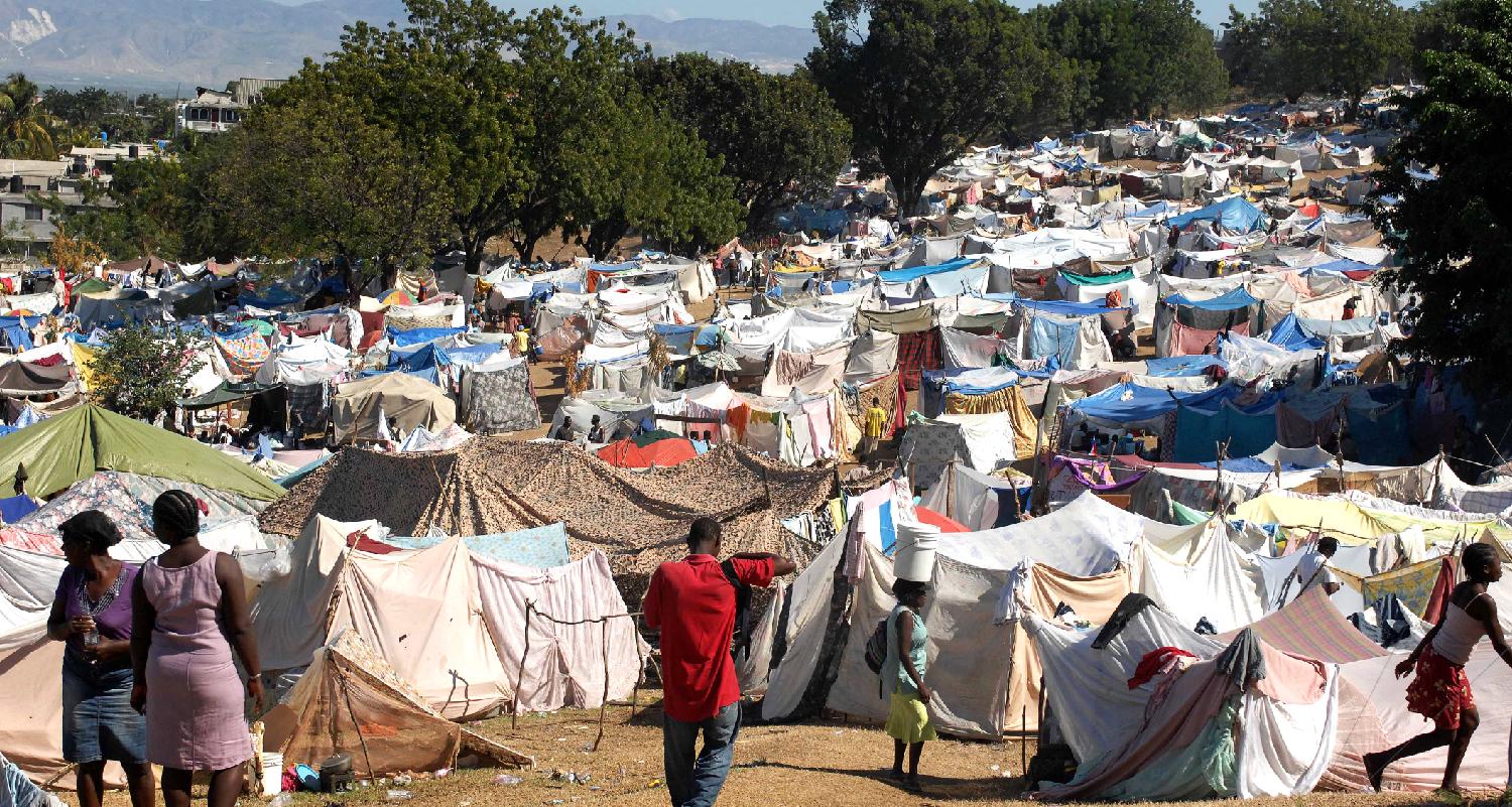 Color photograph of hundreds of tents. The tents are constructed of a variety of cloths of many different colors and are placed very close to one another. A handful of people are visible in the foreground of the image.