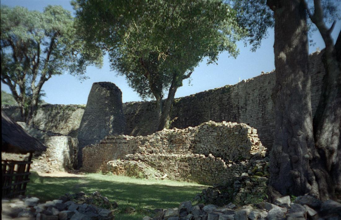 An image of the remains of a stone structure is shown. A tall brick wall is shown in the background with tall trees in front. A broken, shorter brick wall is seen in three rows with a square dark gray stone tower at the left becoming thinner as it goes higher. Grass and gray stones are seen in the forefront. A dark slated object with a triangle top is shown in the bottom left.