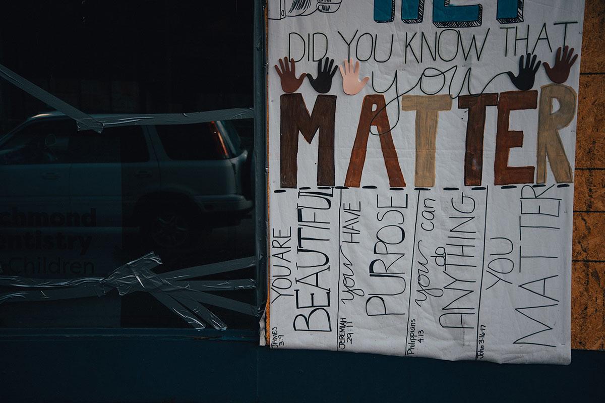 A window is taped and boarded up.  Over the board, a sign hangs reading, “Did You Know That You Matter. You are beautiful. You have Purpose. You can do anything. You matter.”