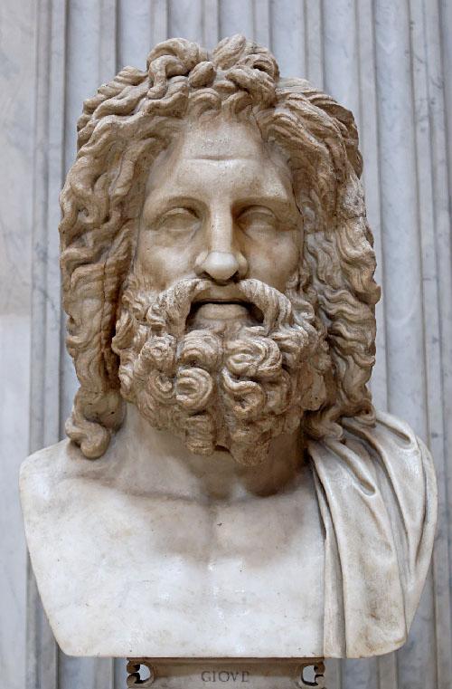 An image of a stone bust of a man is shown on a gray and black marble draped background and pedestal with the letters “GIOVL” carved at the bottom. The man shows shoulder length curly hair and full beard in a light brown color. His eyes are almond shaped and he has a large nose. Over his naked lighter beige left shoulder a cloth is draped.