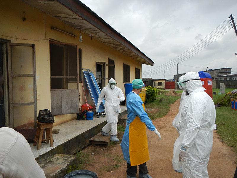 Several people dressed in personal protective equipment outside of a one-story stucco building. Their gear includes face masks, gloves, foot coverings, and goggles.